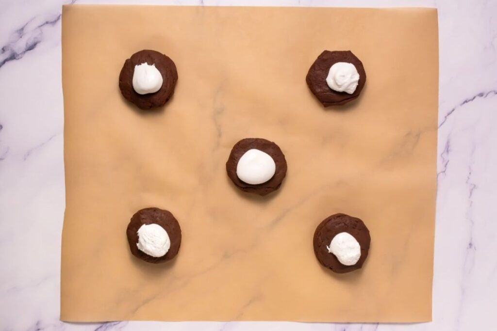 Five chocolate cookie dough rounds with dollops of white cream on top are arranged on a sheet of parchment paper on a marble surface.