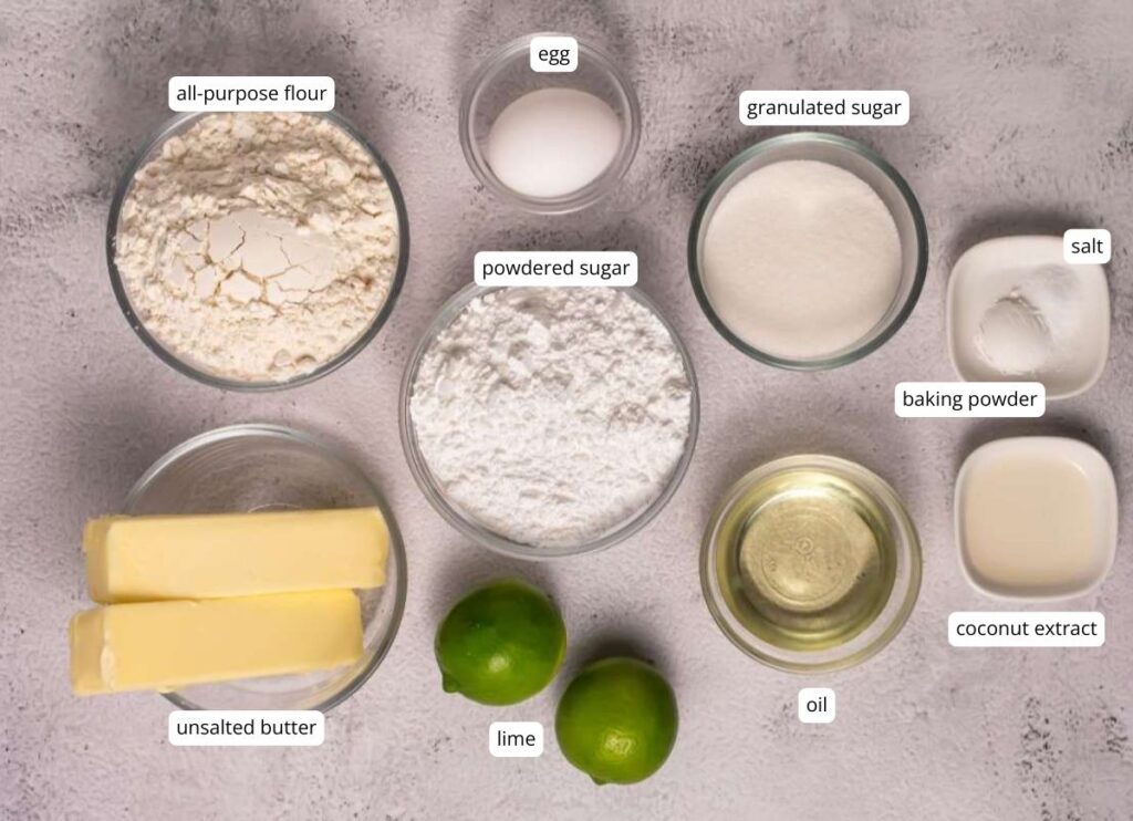 Top-down view of baking ingredients in bowls: all-purpose flour, egg, granulated sugar, powdered sugar, salt, baking powder, coconut extract, oil, unsalted butter, and two limes.