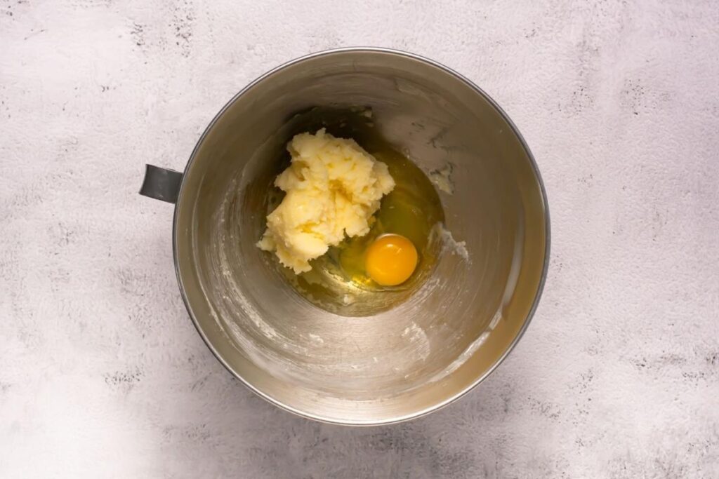 A metal mixing bowl containing creamed butter and sugar with a raw egg cracked on top, ready to be mixed.