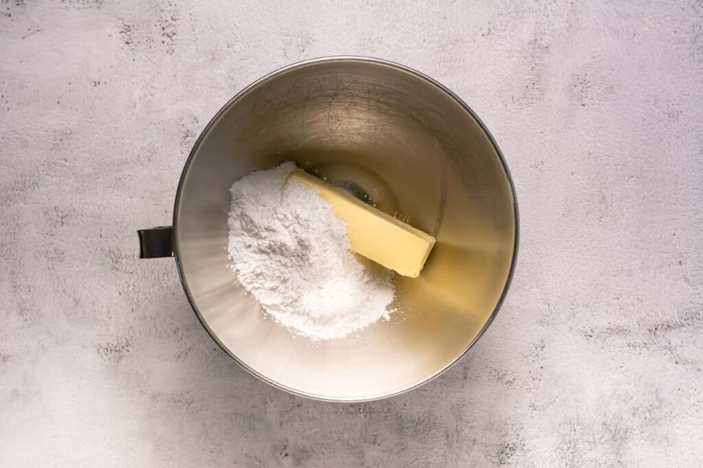 A metal mixing bowl containing a stick of butter and a mound of powdered sugar on a light gray textured surface.