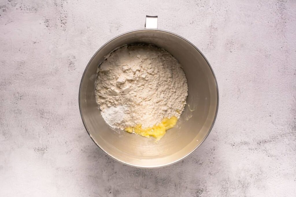 A metal mixing bowl containing flour, sugar, and softened butter on a light-colored textured surface.