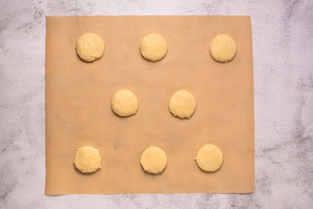Nine dough balls are evenly spaced on a sheet of parchment paper, ready for baking, set on a light-colored countertop.