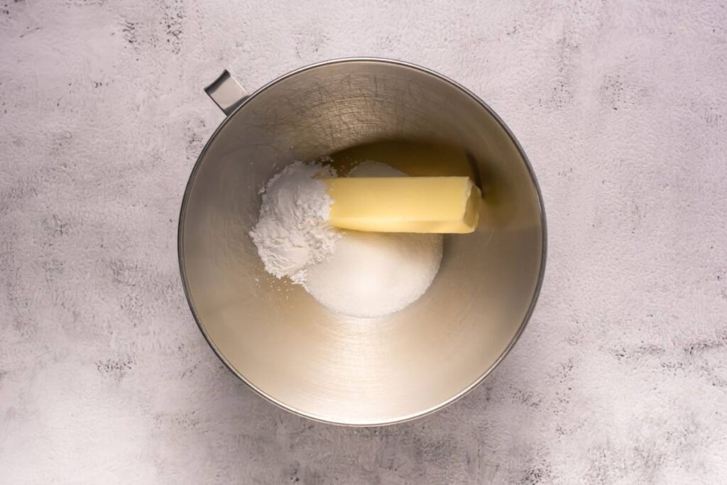 A metal mixing bowl contains a stick of butter, a pile of white granulated sugar, and a pile of powdered sugar on a light gray surface.