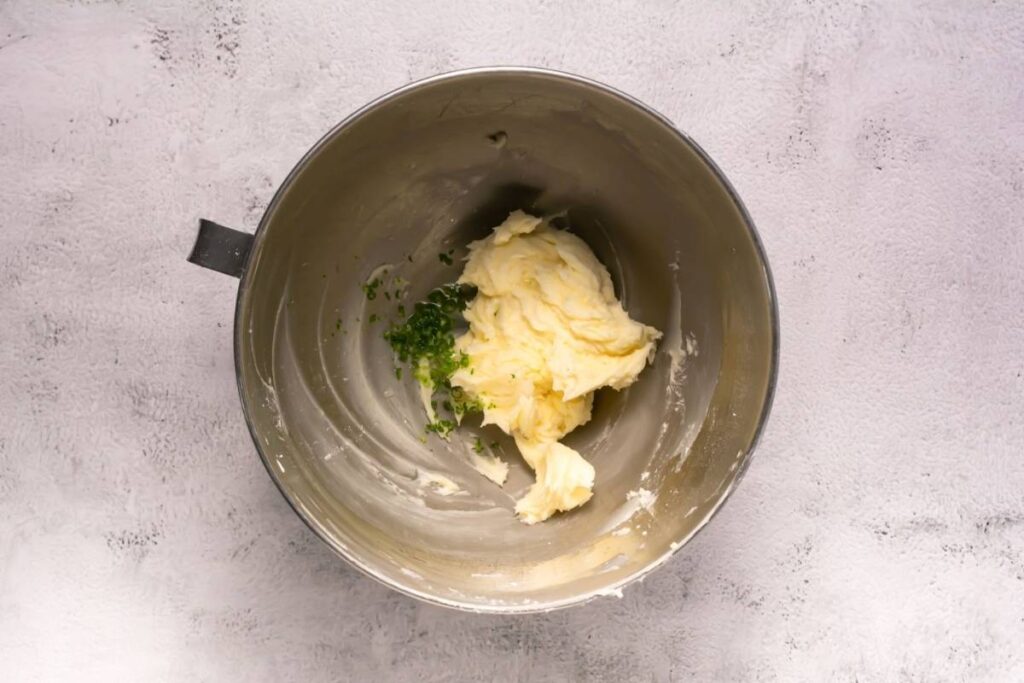 A mixing bowl containing whipped butter and chopped green herbs on a light textured surface.
