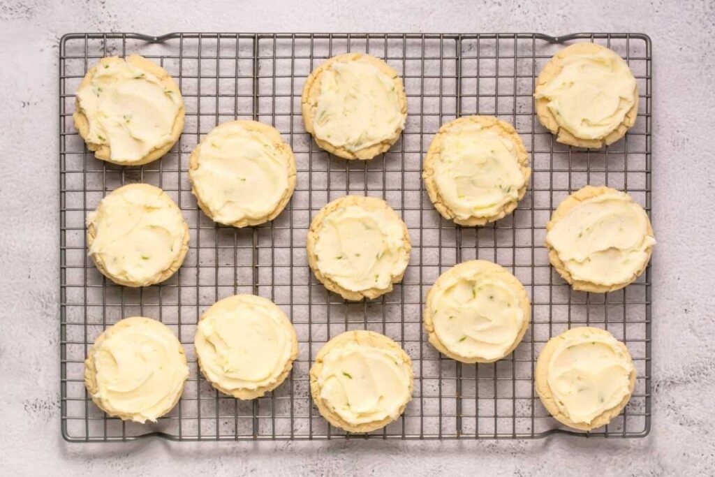 Thirteen round cookies with pale yellow frosting are arranged on a cooling rack over a light-colored surface.
