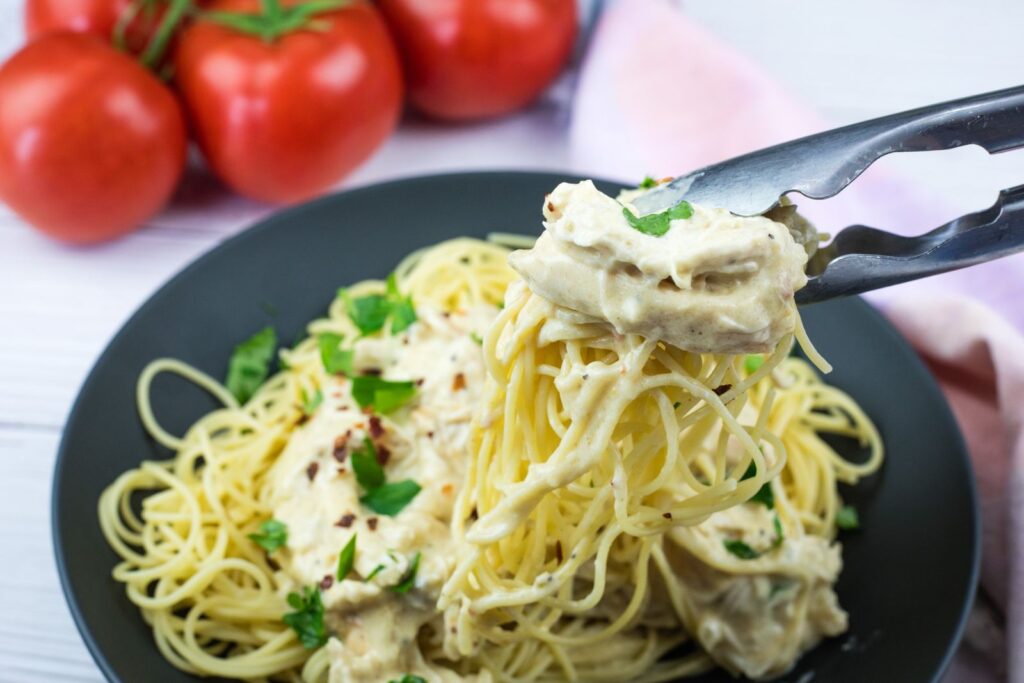 A close-up of tongs lifting creamy chicken and spaghetti from a black plate, garnished with parsley, with fresh tomatoes in the background.