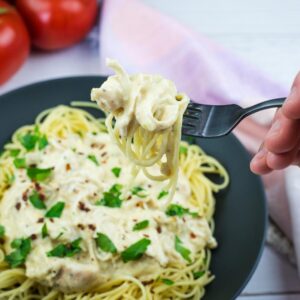 A hand holding a fork with a twirl of spaghetti above a plate of pasta topped with creamy sauce and parsley; tomatoes are in the background.