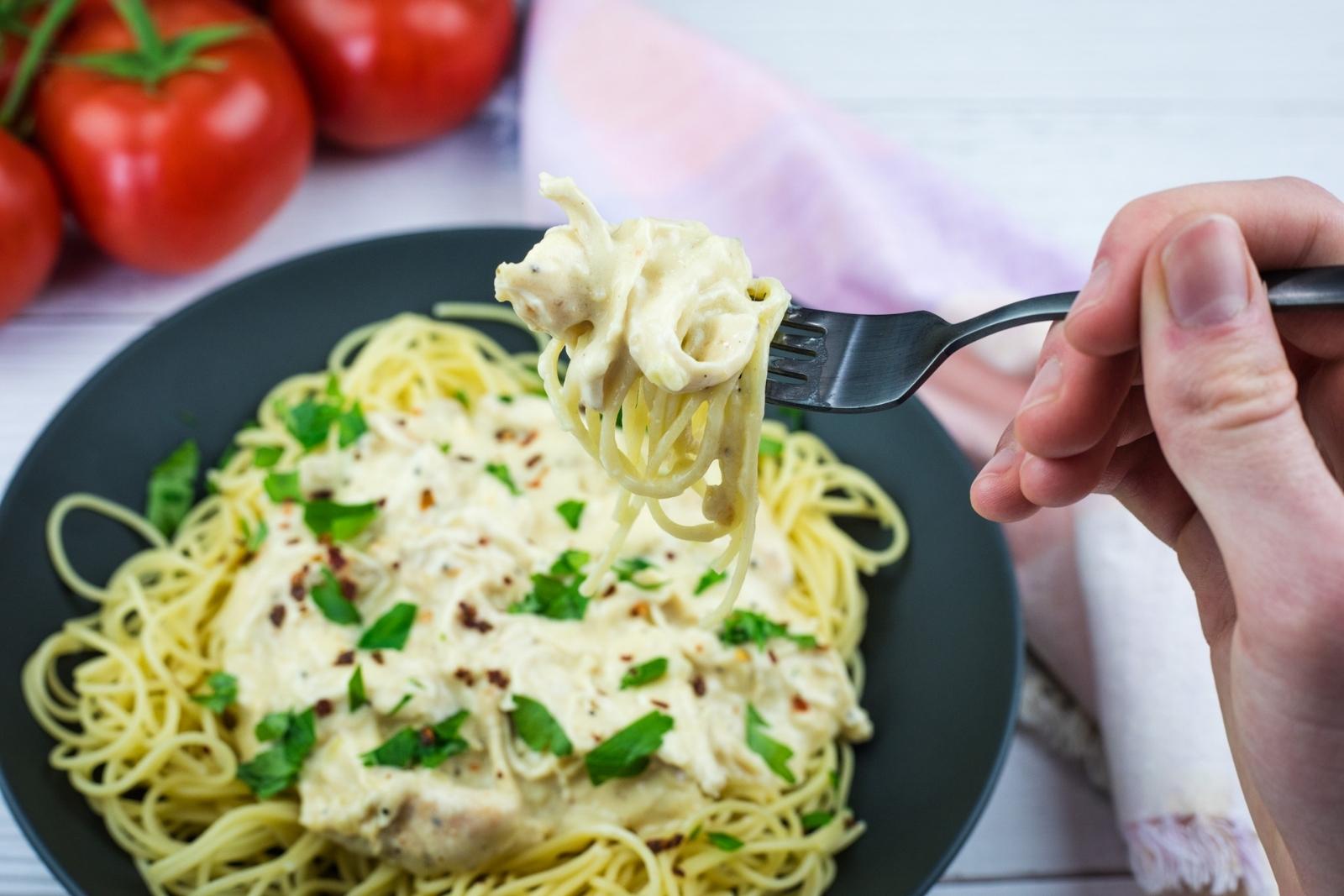 A hand holding a fork with a twirl of spaghetti above a plate of pasta topped with creamy sauce and parsley; tomatoes are in the background.
