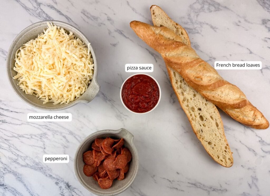 Bowls of shredded mozzarella cheese, sliced pepperoni, a cup of pizza sauce, and two French bread loaves are arranged on a marble surface.