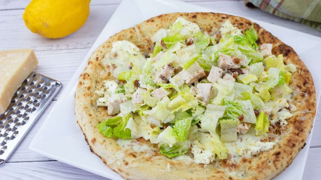 A pizza topped with pieces of lettuce, chicken, grated cheese, and dressing, served on a white plate with a lemon and cheese wedge nearby.