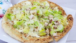 A pizza topped with chopped romaine lettuce, grilled chicken pieces, and a creamy dressing, served on a white plate.