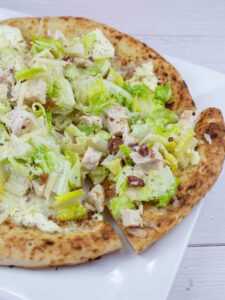 A close-up of a pizza topped with chopped lettuce, chicken pieces, bacon bits, cheese, and a creamy dressing on a white plate.