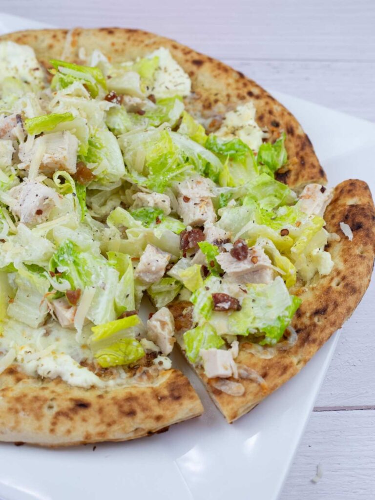 A close-up of a pizza topped with chopped lettuce, chicken pieces, bacon bits, cheese, and a creamy dressing on a white plate.