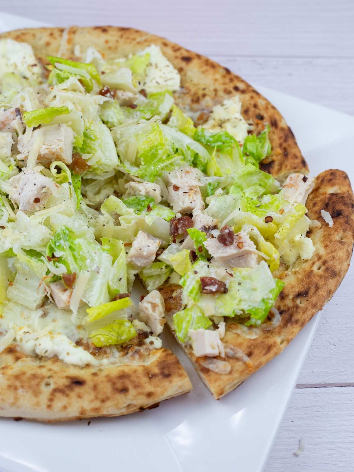 A close-up of a pizza topped with chopped lettuce, chicken pieces, bacon bits, cheese, and a creamy dressing on a white plate.