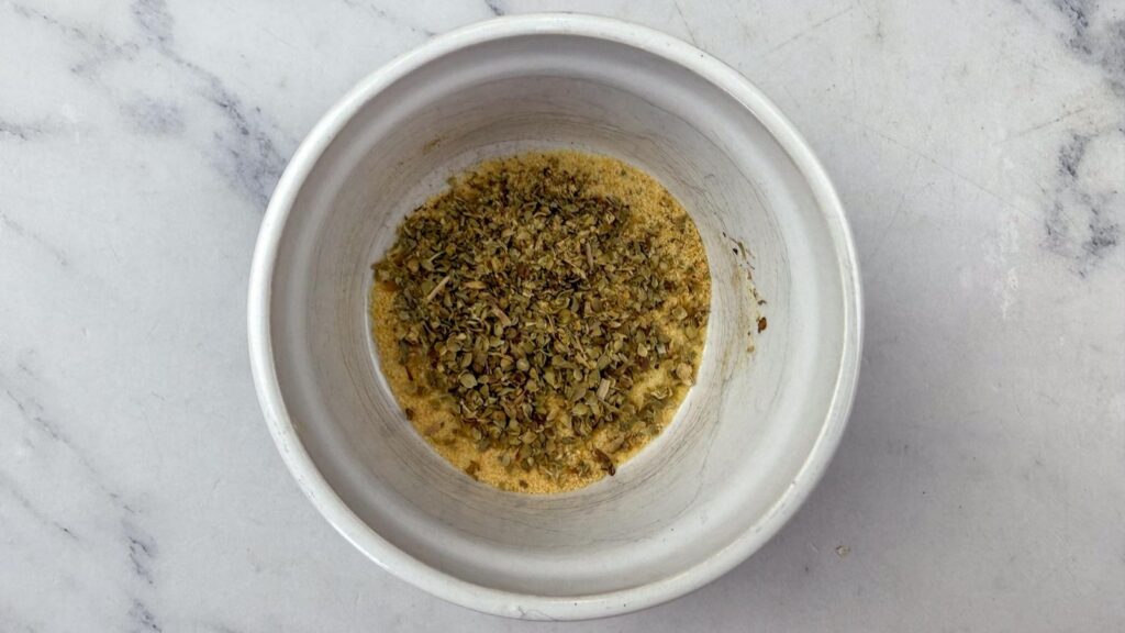 A white bowl on a marble surface containing a mixture of dried herbs and a yellow powder.
