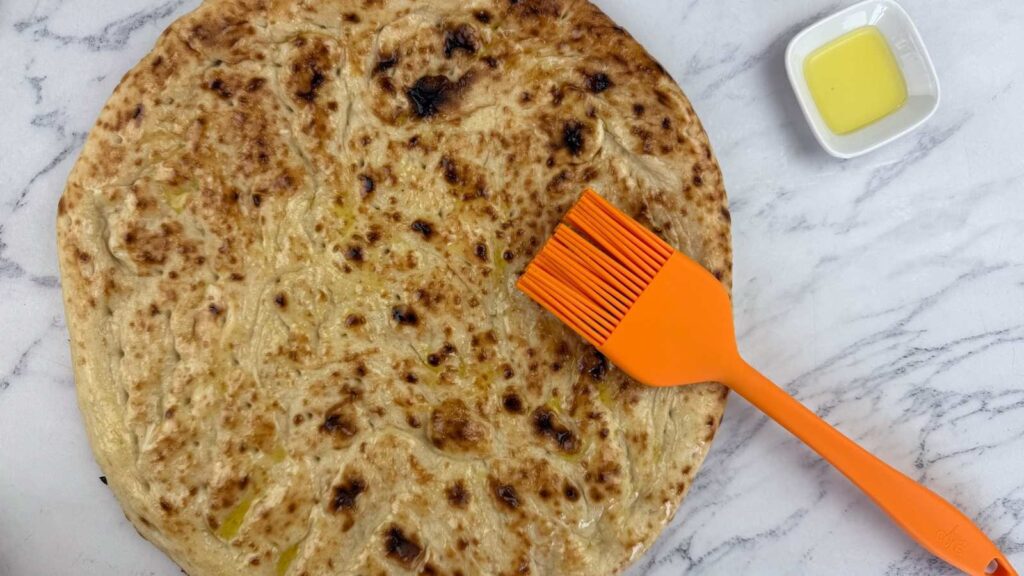 A round flatbread on a marble surface, brushed with oil using an orange silicone brush; a small dish of oil is beside it.