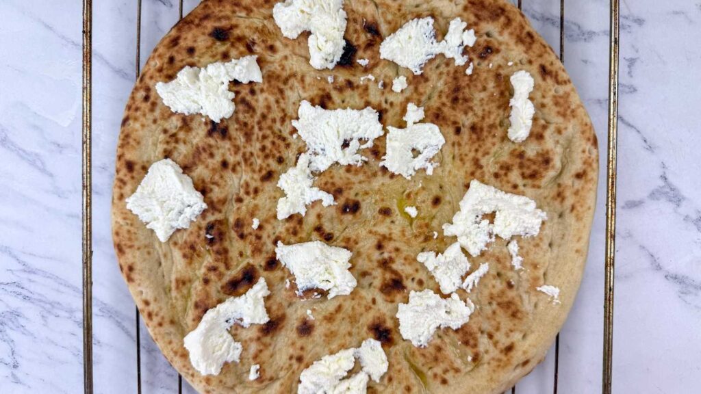 A round flatbread topped with uneven dollops of white ricotta cheese, placed on a metal cooling rack over a marble surface.