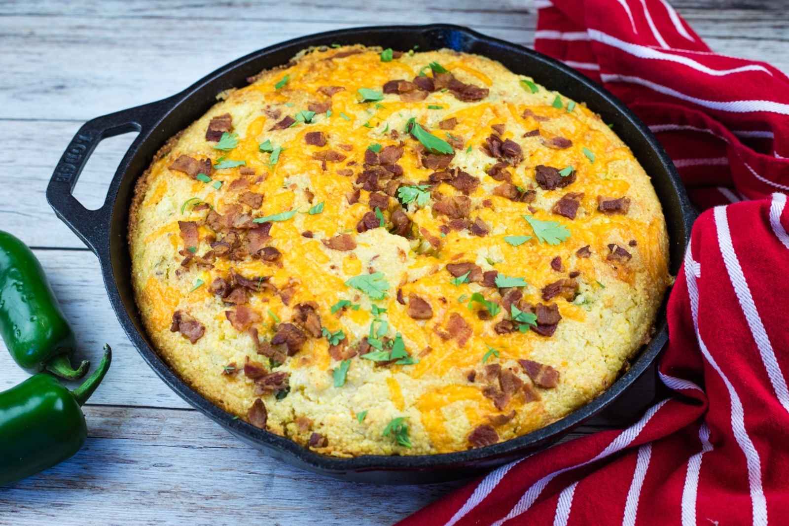A cast-iron skillet filled with baked cornbread topped with chopped bacon and herbs, placed next to a red striped kitchen towel.
