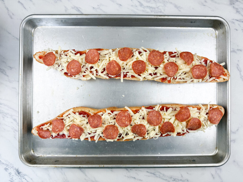 Two pieces of French bread on a baking sheet topped with pizza sauce, shredded cheese, and pepperoni slices, ready to be baked.