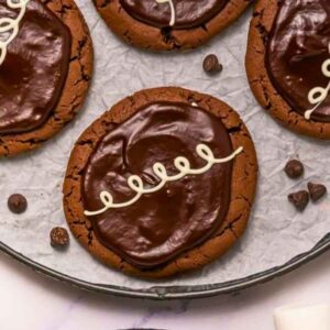 Four chocolate cookies with glossy chocolate icing and a white icing swirl pattern are arranged on a round, parchment-lined tray. Chocolate chips and marshmallows are nearby.