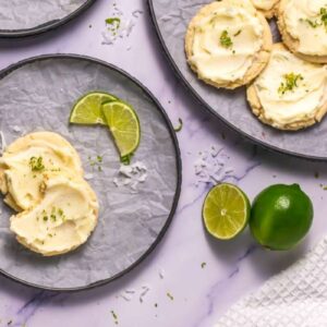 Frosted cookies with lime zest on plates, served with lime slices and shredded coconut on a white marble surface.