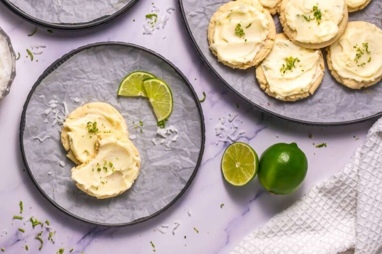 Frosted cookies with lime zest on plates, served with lime slices and shredded coconut on a white marble surface.