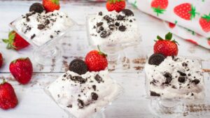 Four glass dessert cups filled with cookies and cream mousse, topped with mini chocolate cookies and strawberries, placed on a white wooden surface with loose strawberries around.