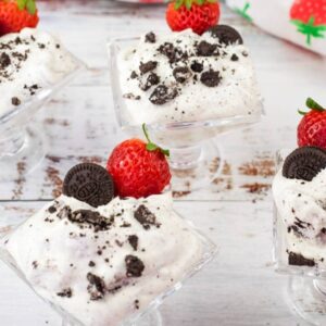 Four glass dessert cups filled with cookies and cream mousse, topped with mini chocolate cookies and strawberries, placed on a white wooden surface with loose strawberries around.
