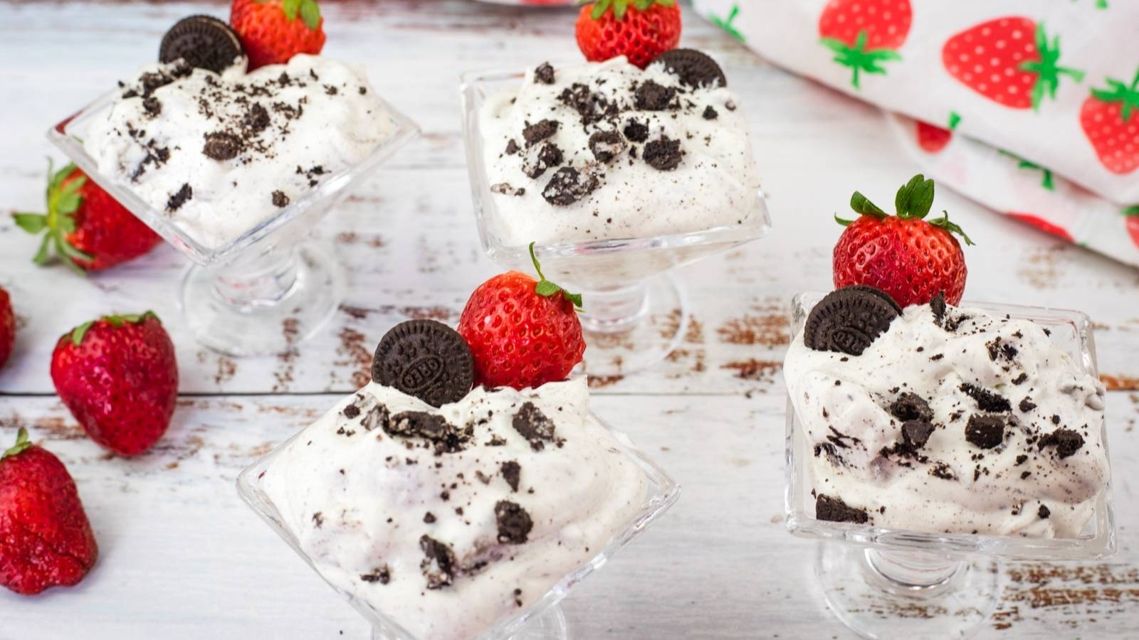 Four glass dessert cups filled with cookies and cream mousse, topped with mini chocolate cookies and strawberries, placed on a white wooden surface with loose strawberries around.