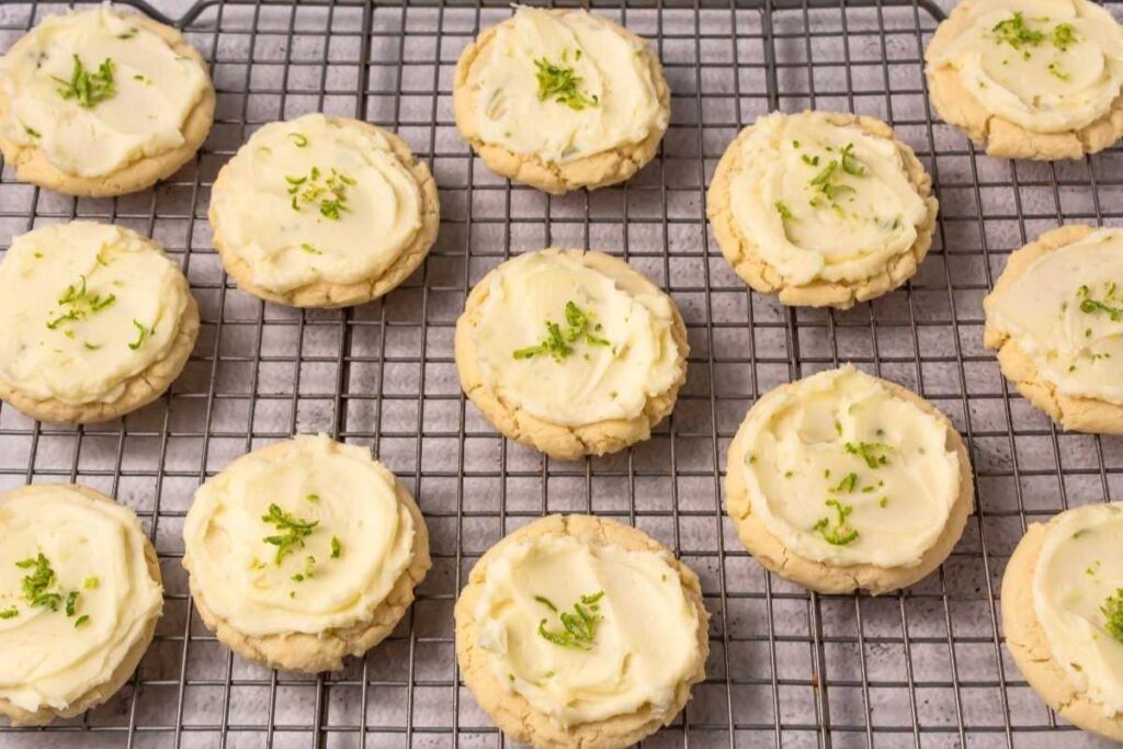 Cookies with white frosting and green zest garnish are arranged on a wire cooling rack.