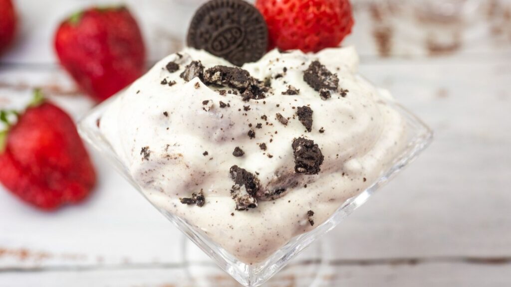 A glass cup filled with cookies and cream mousse, topped with a mini chocolate sandwich cookie and surrounded by fresh strawberries.