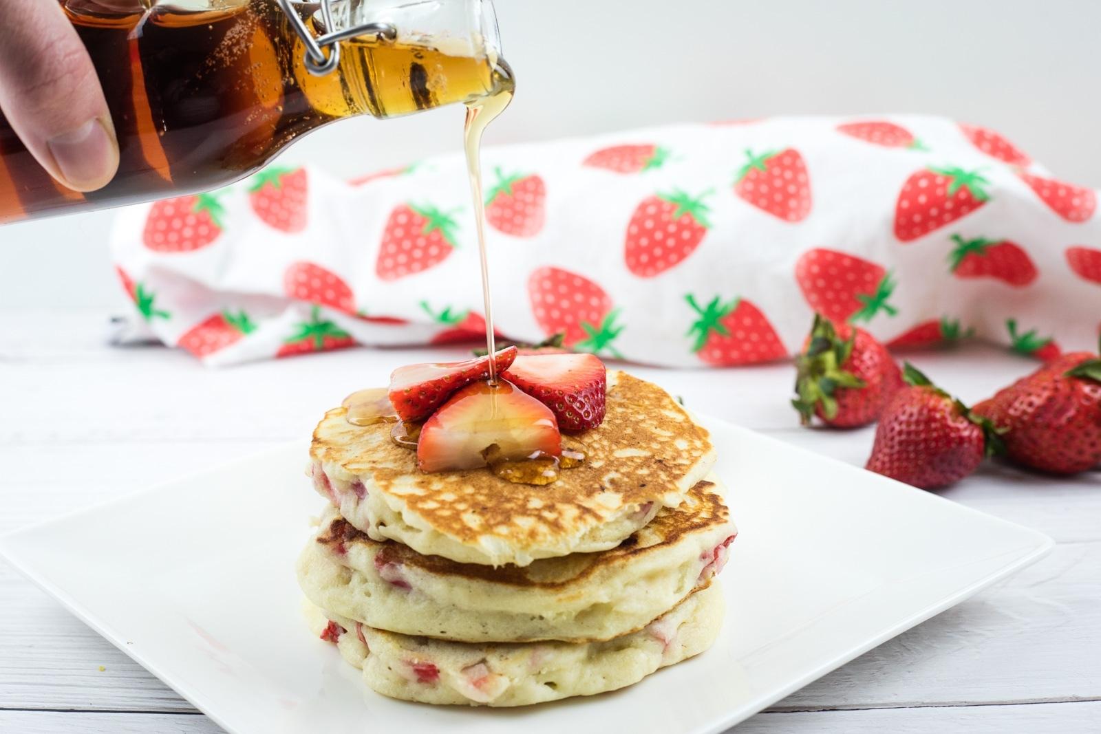 A stack of pancakes with sliced strawberries on top is being drizzled with syrup, with whole strawberries and a strawberry-patterned cloth in the background.