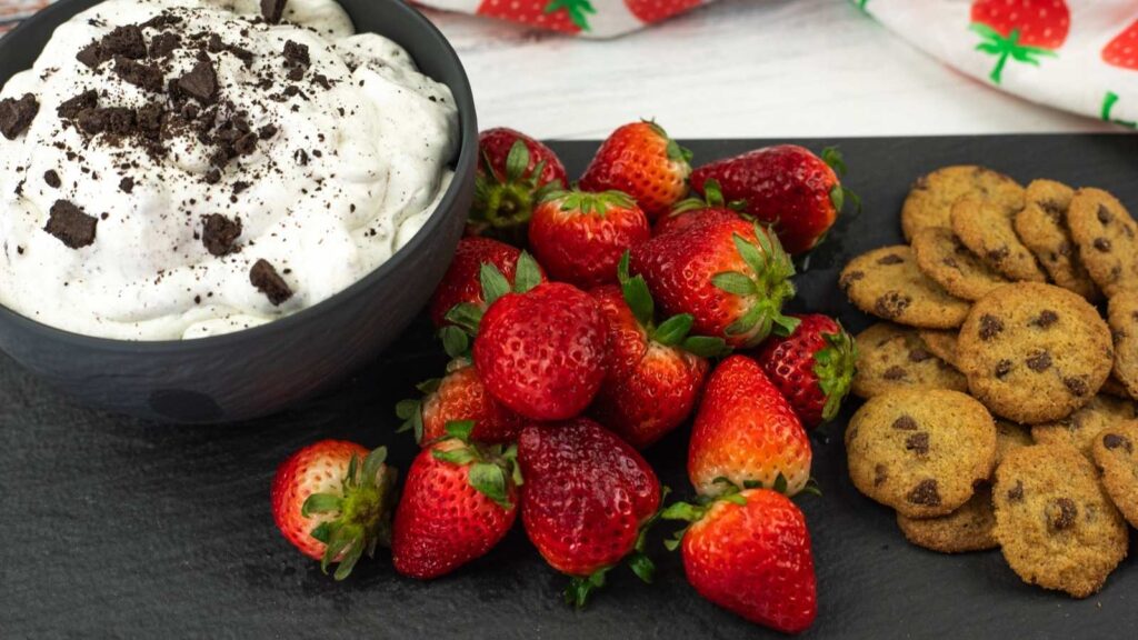 A bowl of whipped cream topped with cookie crumbs, a pile of fresh strawberries, and a group of mini chocolate chip cookies on a black serving tray.