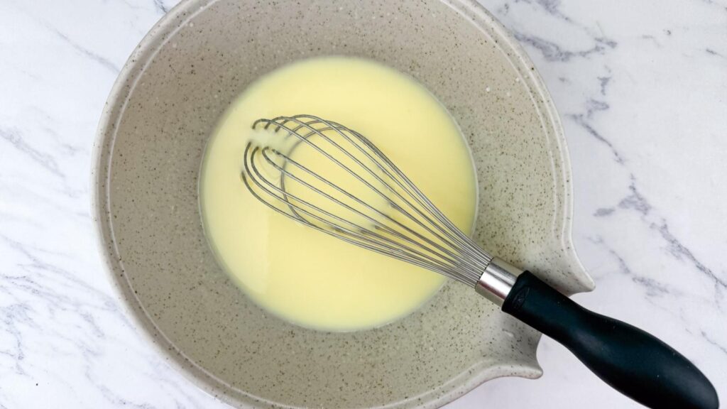 A metal whisk rests in a beige mixing bowl containing a pale yellow liquid, placed on a white marble surface.
