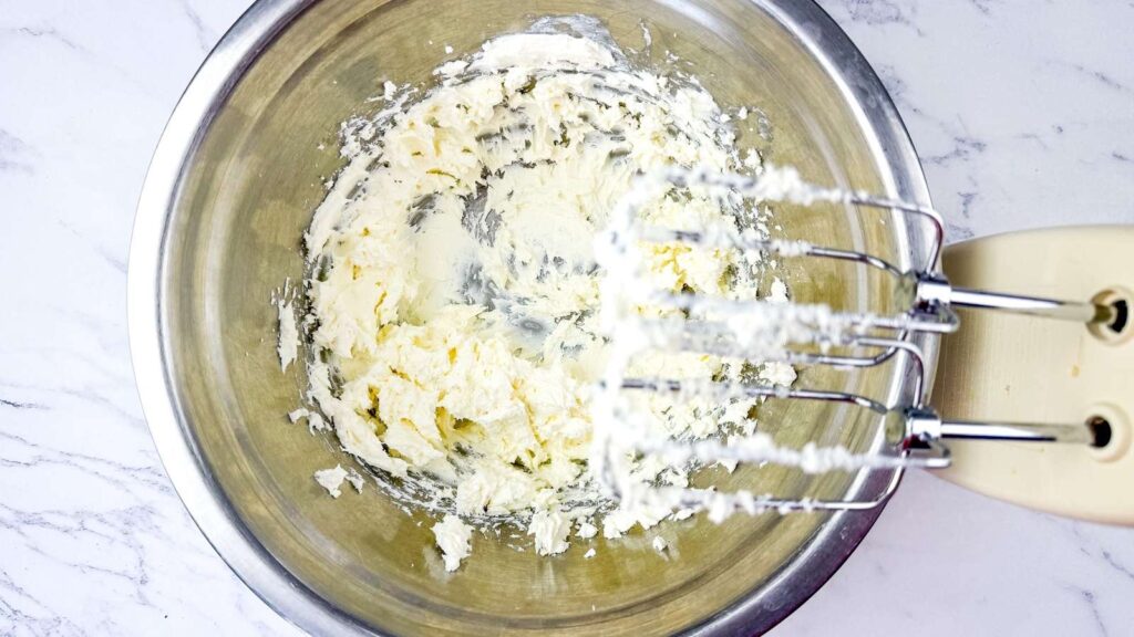 A metal mixing bowl with creamed butter and sugar, next to handheld electric mixer beaters covered in the mixture, on a marble countertop.