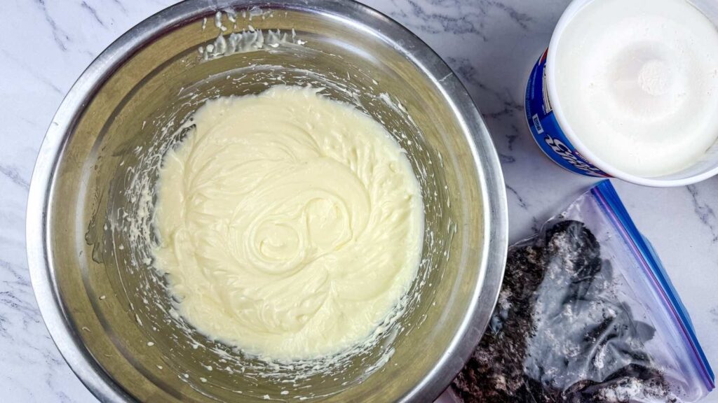 A stainless steel bowl with a creamy, white mixture next to a tub of whipped topping and a zip-top bag containing crushed cookies on a marble countertop.