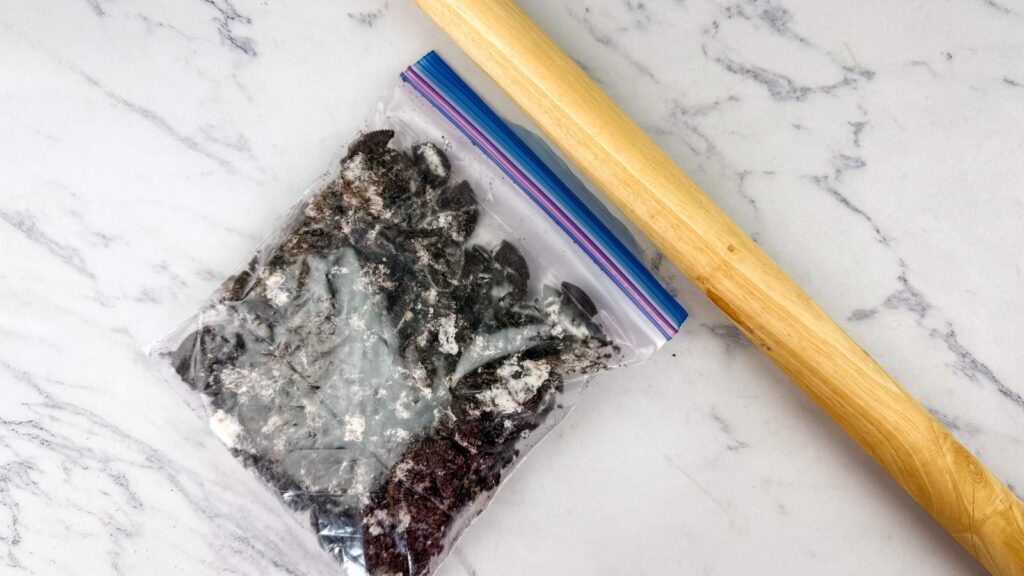 A plastic bag filled with crushed chocolate cookies lies on a marble surface next to a wooden rolling pin.