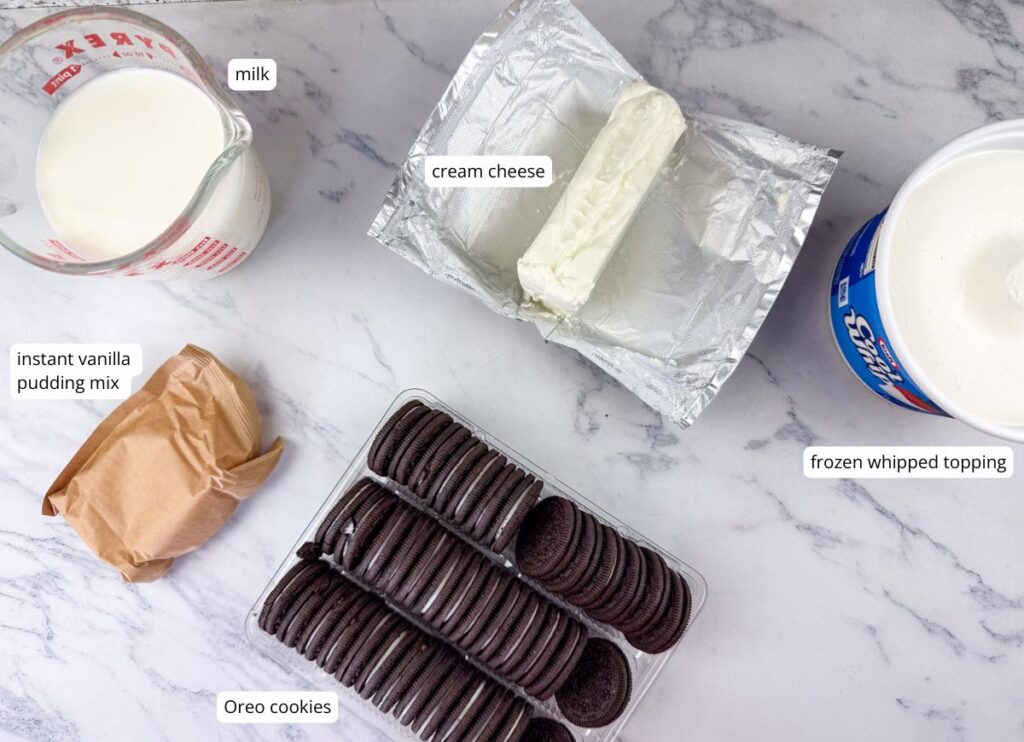 Ingredients on a marble surface: a measuring cup of milk, cream cheese, frozen whipped topping, instant vanilla pudding mix, and a tray of Oreo cookies.