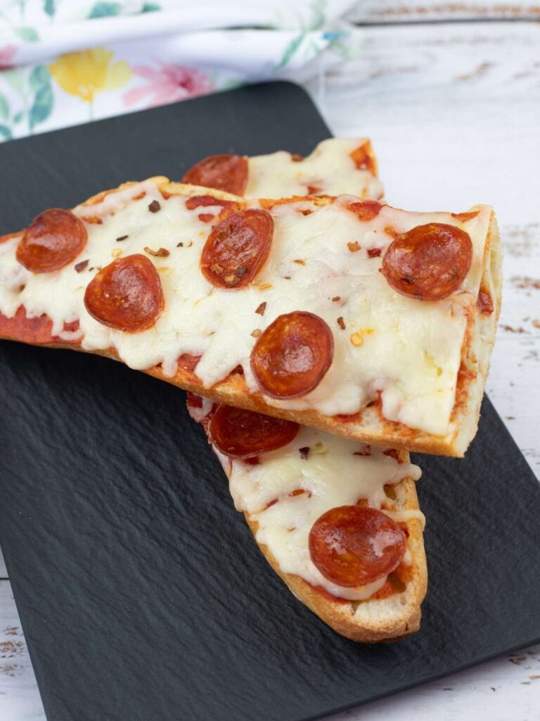 Two slices of French bread pizza topped with melted cheese and pepperoni are placed on a black slate serving board.