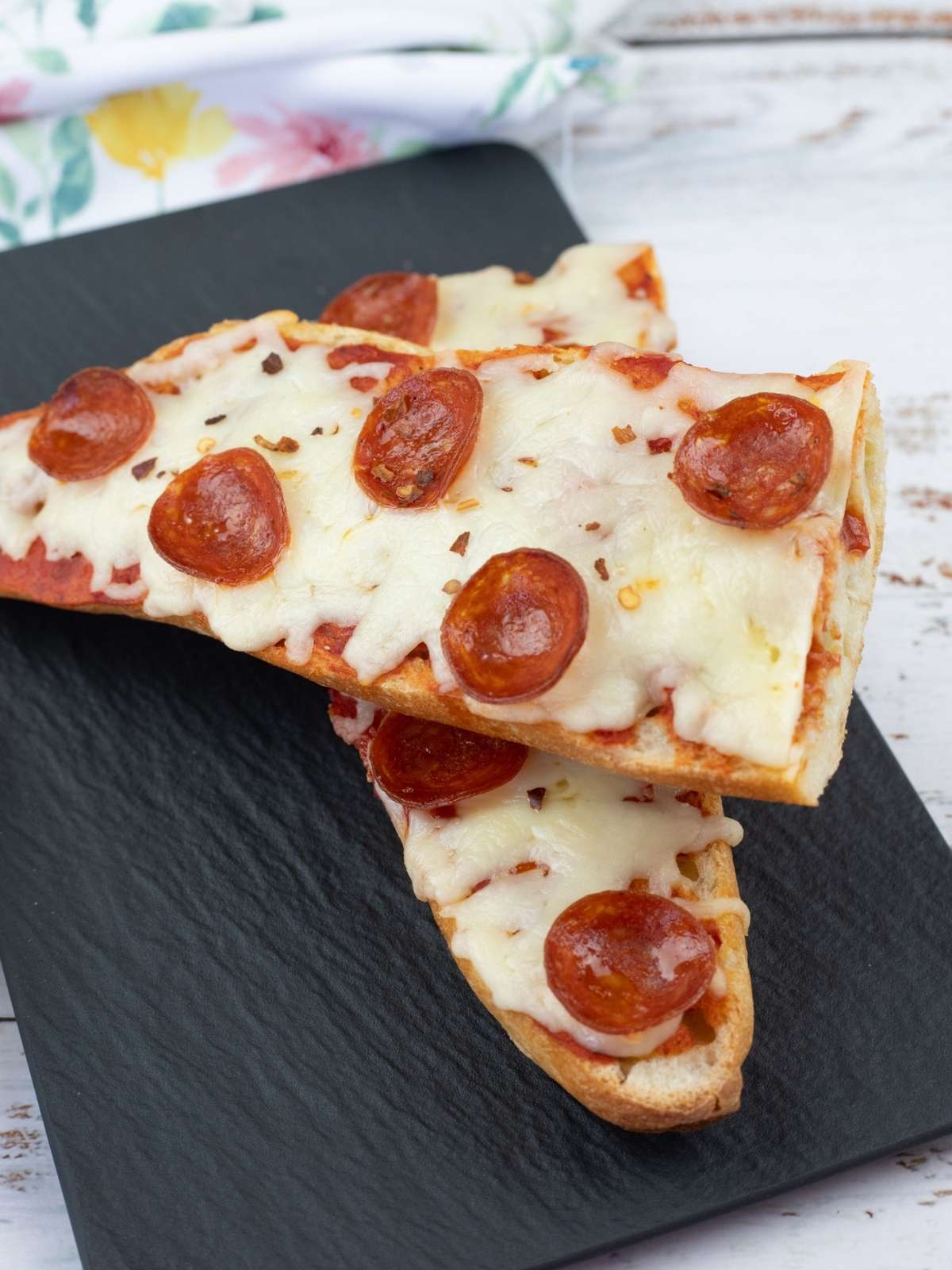 Two slices of French bread pizza topped with melted cheese and pepperoni are placed on a black slate serving board.