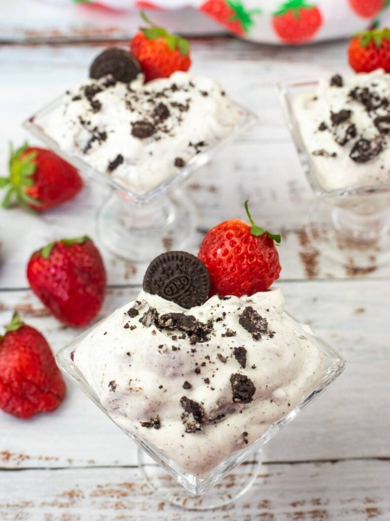 Three glass dishes filled with cookies and cream dessert, each topped with a mini chocolate cookie and a fresh strawberry, are arranged on a white wooden surface.