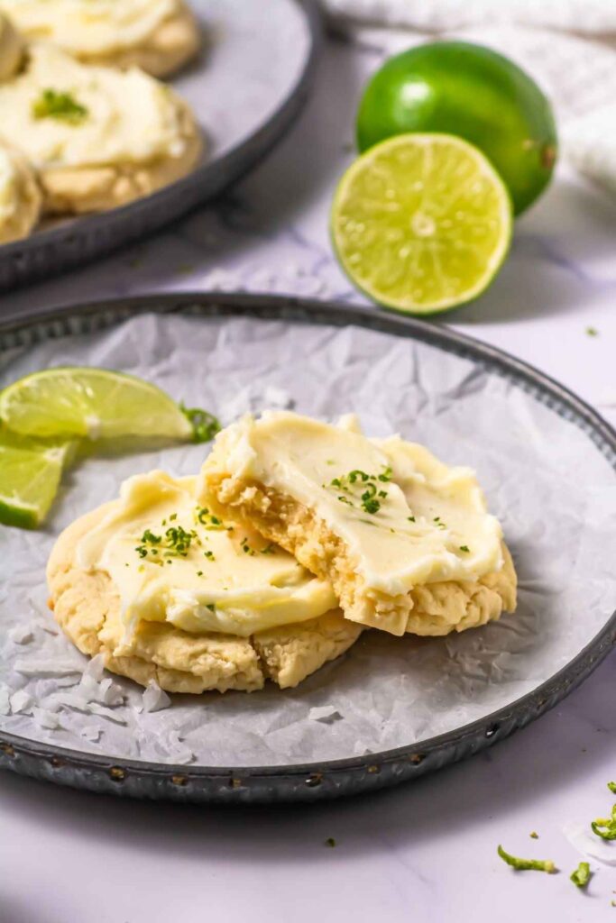Two frosted lime cookies on a plate, one broken in half, garnished with lime zest and a lime wedge, with whole and halved limes in the background.