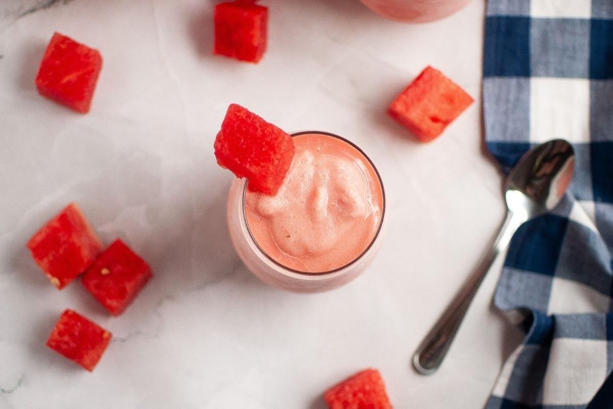 A glass of pink watermelon smoothie topped with a watermelon cube, surrounded by watermelon pieces, a spoon, and a blue checkered cloth on a white surface.