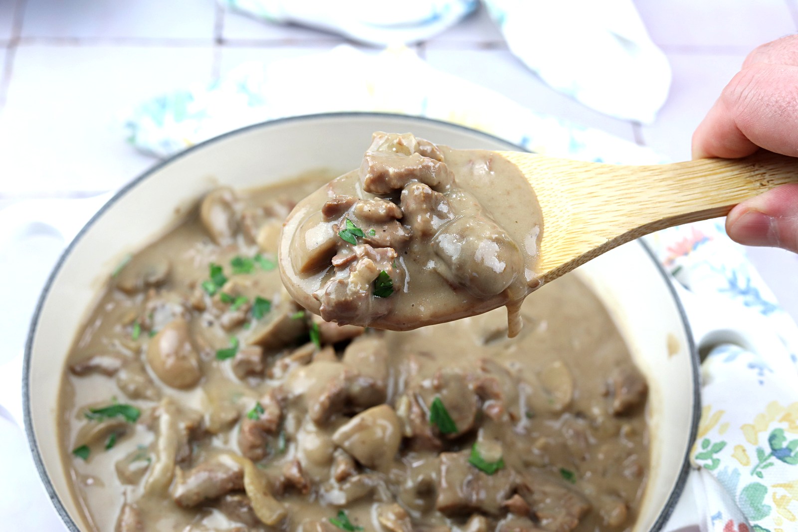 A wooden spoon holding a serving of creamy beef and mushroom stew over a pot filled with the same dish.