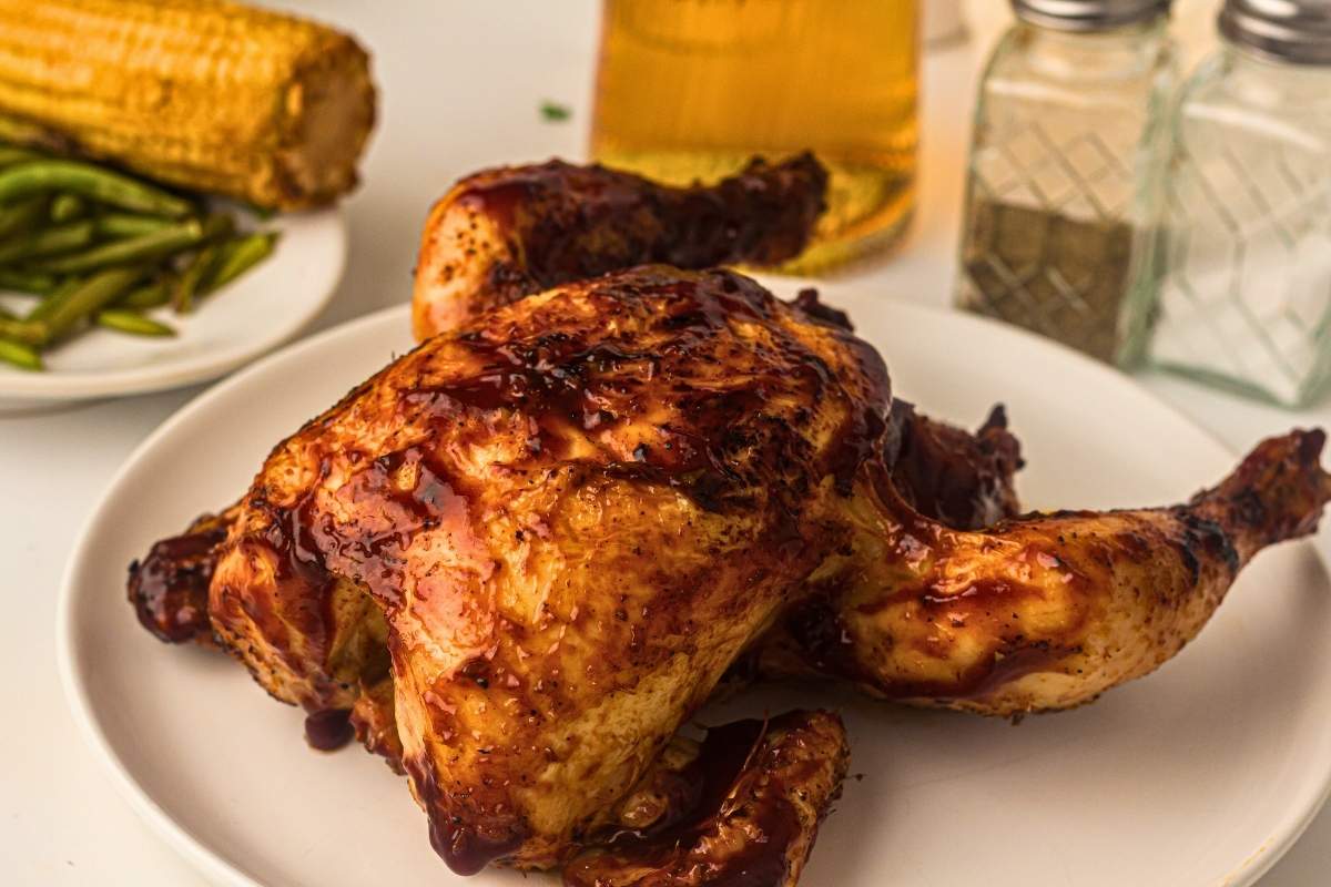 A roasted chicken with barbecue sauce on a white plate, with green beans, corn on the cob, a glass of beer, and salt and pepper shakers in the background.