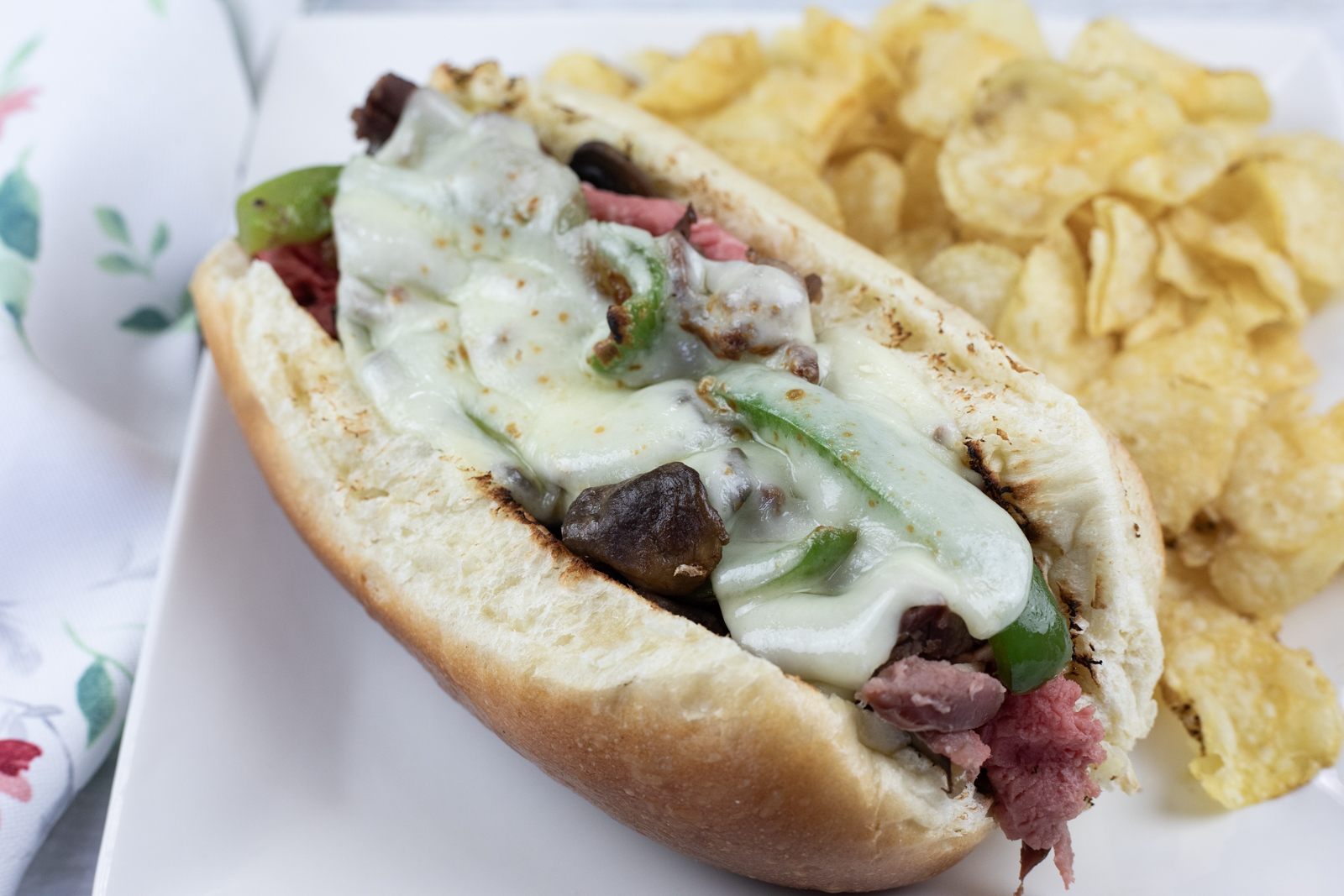 A Philly cheesesteak sandwich with melted cheese, green peppers, and onions in a hoagie roll, served with potato chips on a white plate.