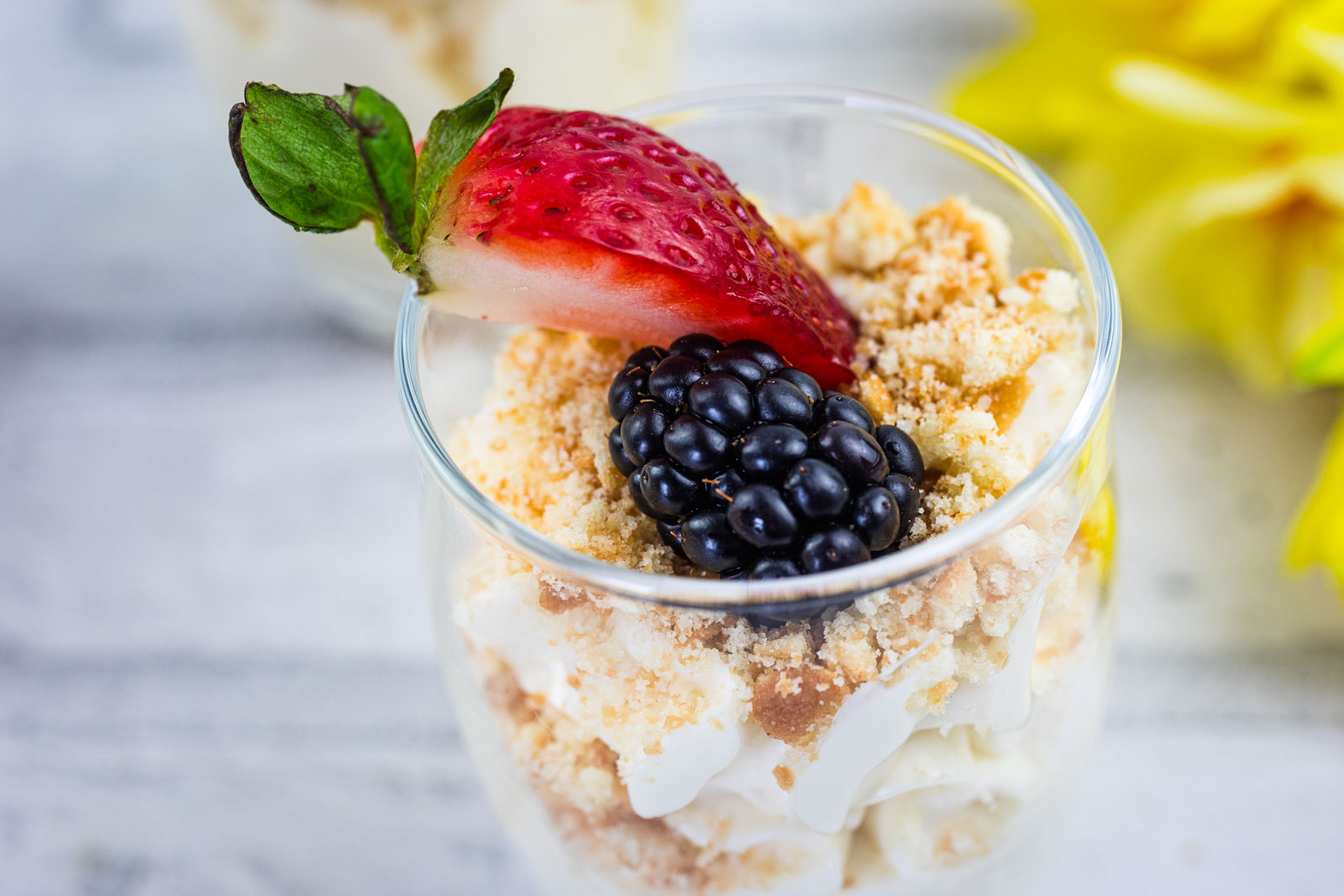 A dessert cup filled with crumbled cookies, cream, topped with a sliced strawberry and a blackberry, set on a light surface with yellow flowers in the background.