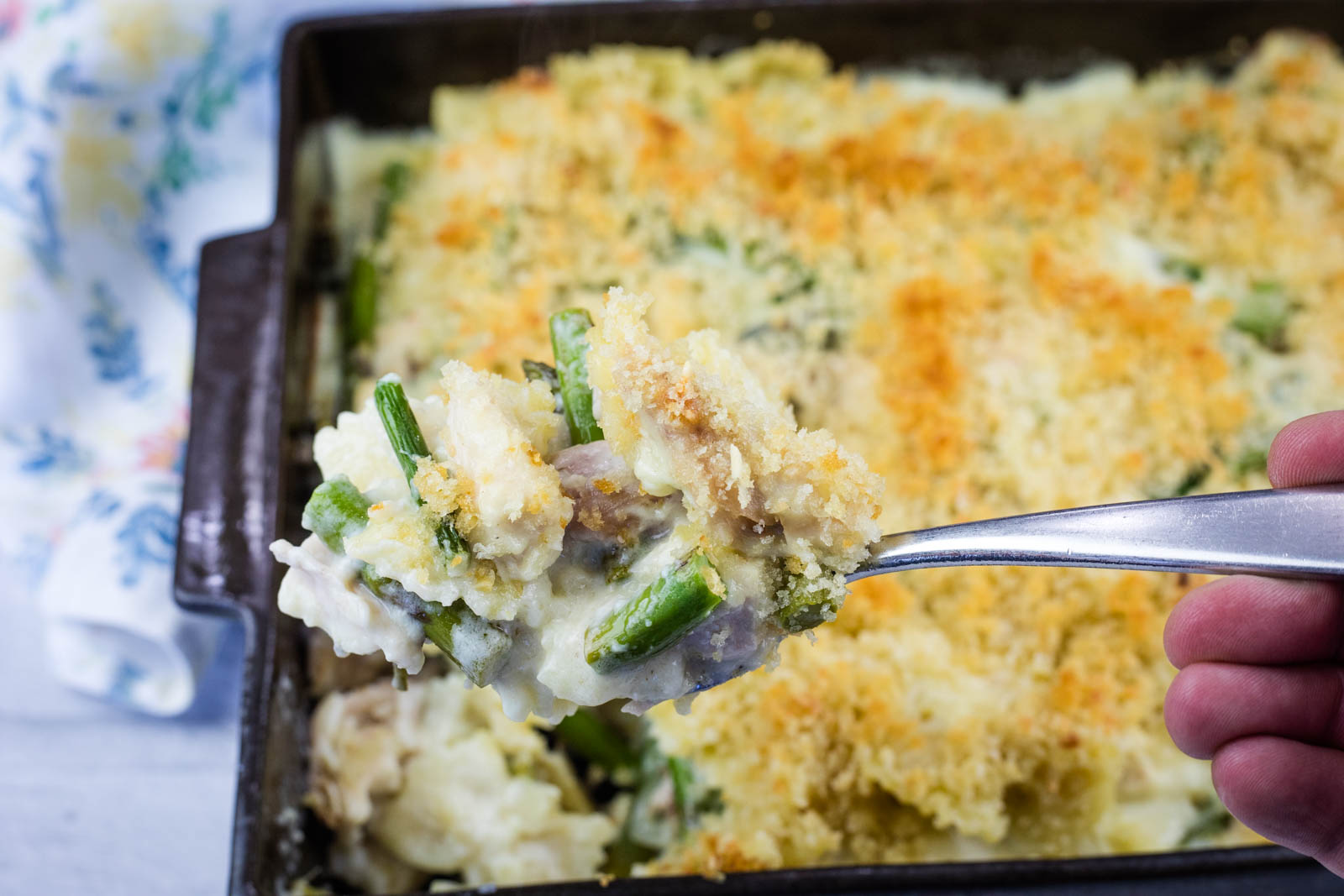 A hand holds a spoonful of Gruyère Chicken Asparagus Pasta casserole topped with breadcrumbs over a baking dish filled with the same casserole.