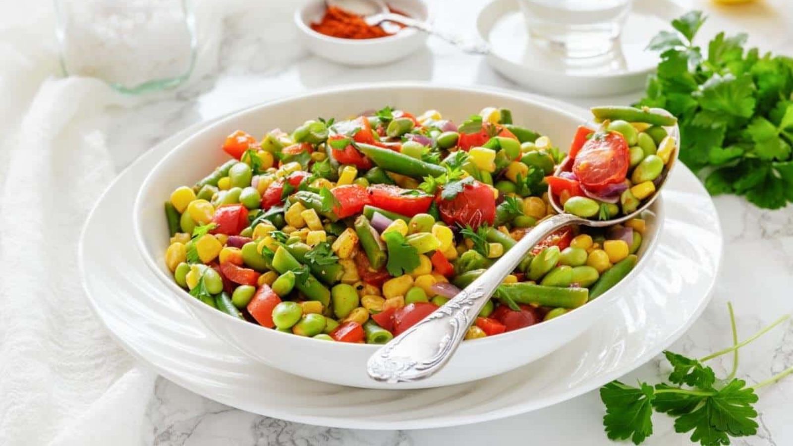 A bowl of colorful bean salad with corn, tomatoes, green beans, and herbs, served with a spoon on a white table.