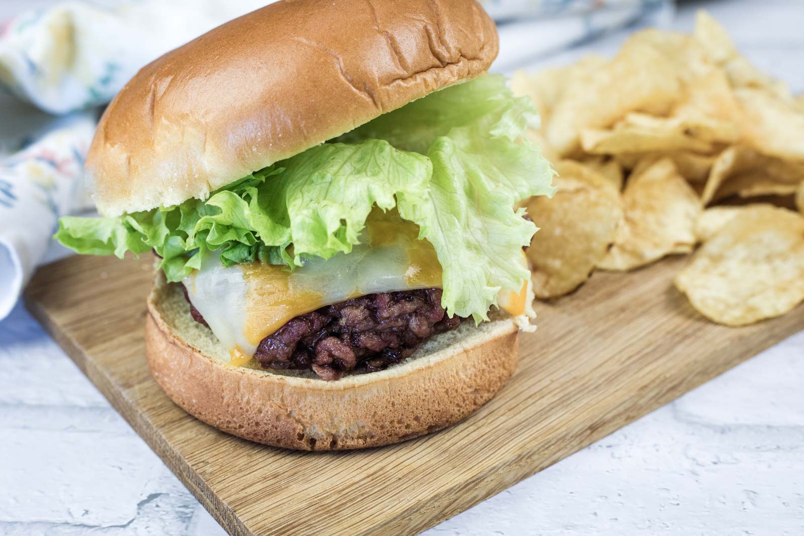 A cheeseburger with lettuce and cheese on a bun sits on a wooden board next to a pile of potato chips.